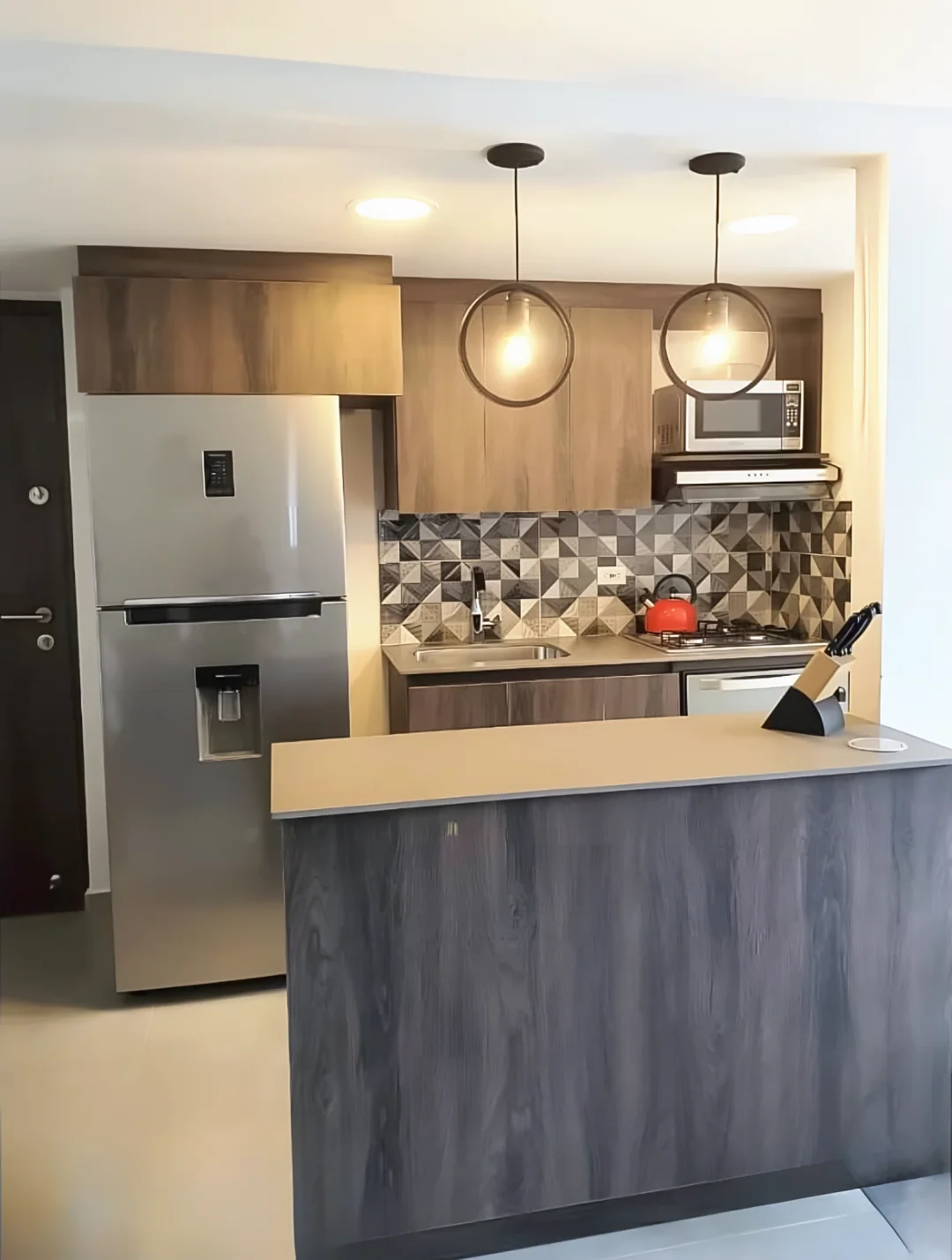 Cocina abierta con barra en madera oscura, alacenas en madera, lámparas colgantes y enchape geométrico