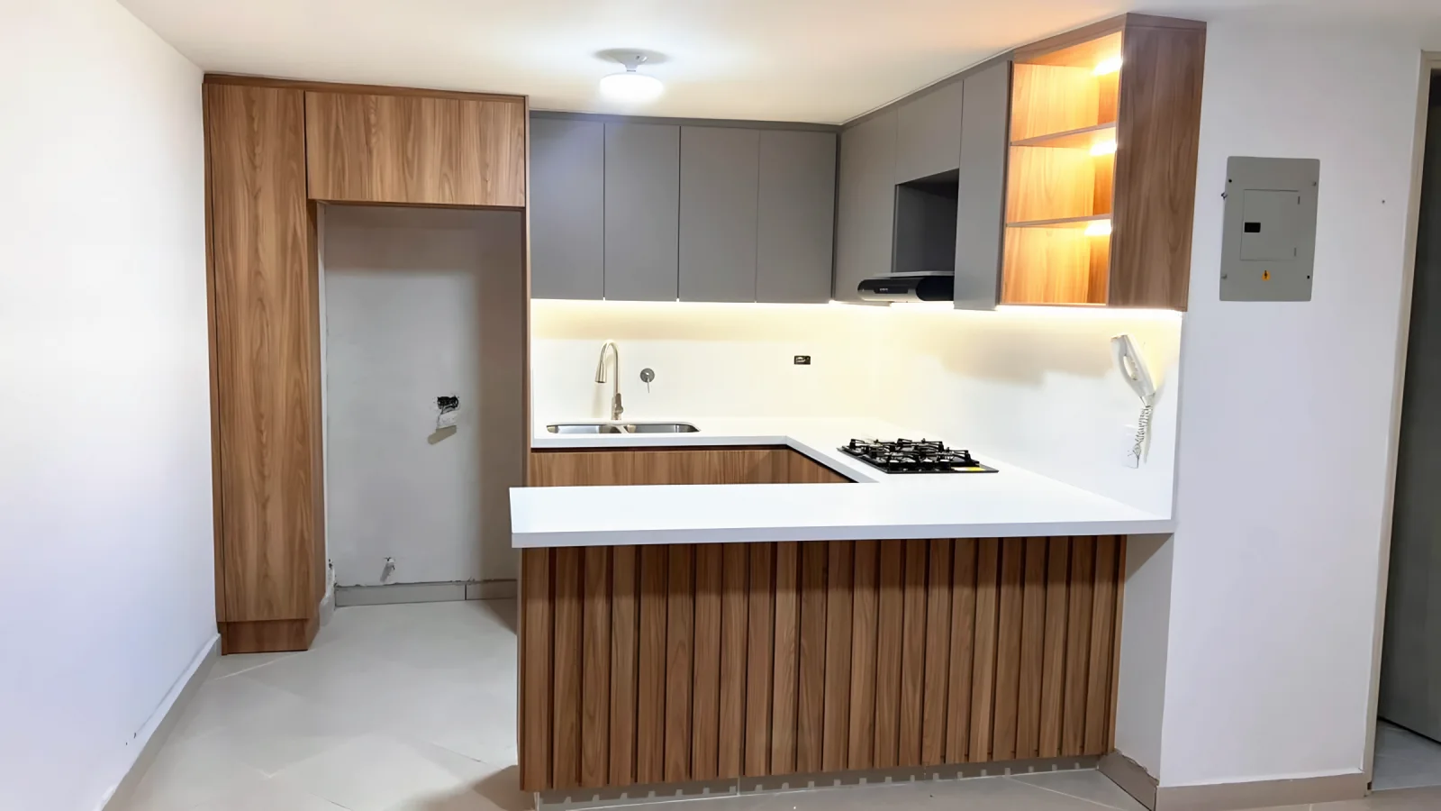 Vista frontal de la cocina en U con barra en listones de madera, mesón blanco y torre alta junto a la entrada